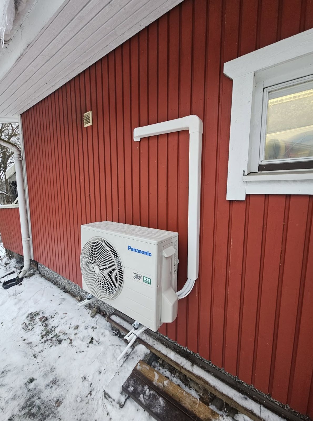 Panasonic-ilmalämpöpumpun ulkoyksikkö asennettuna punaisen omakotitalon seinustalle talvella siistillä putkikanavalla